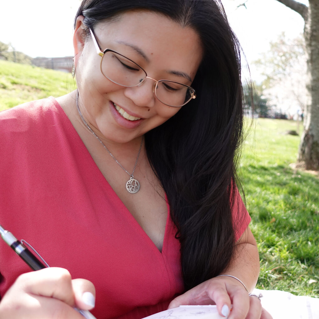 Photo of Melissa sketching under the cherry blossom trees in a pink dress, and wearing her custom US Capitol cherry blossom necklace.